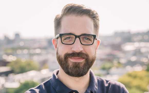 A man with brown hair, a beard, and black glasses smiling against a blurred urban background.
