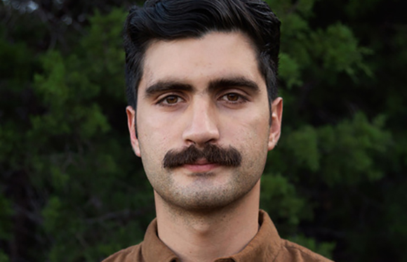 Portrait of a man with dark hair, brown eyes, and a thick mustache against a background of green foliage.