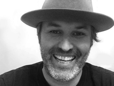 Black and white portrait of a smiling man with a beard wearing a wide-brimmed hat.