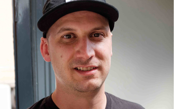 A man with short facial hair wearing a black baseball cap and a black t-shirt.