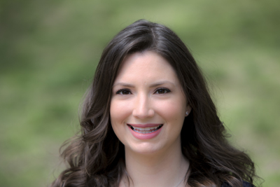 A smiling woman with long, wavy brown hair looks toward the camera against a blurred green background.