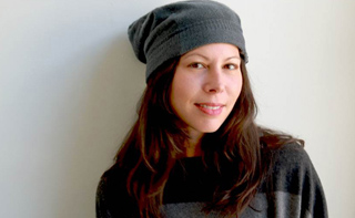 A woman with long dark hair wearing a grey beanie and a dark patterned sweater against a light background.