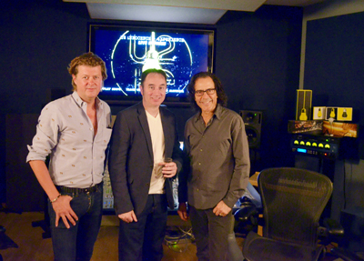 Three men standing side by side in a dark music studio in front of a glowing screen.