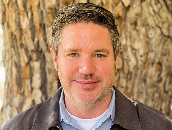 A man with short brown hair and light stubble smiling in front of a textured tree trunk.