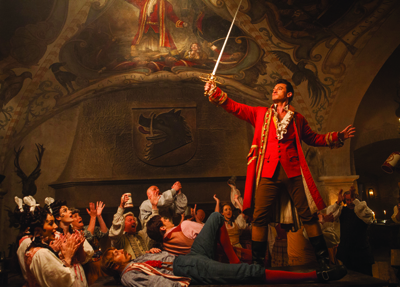 A man in a red coat stands with a sword raised, surrounded by a cheering crowd in a dimly lit hall.