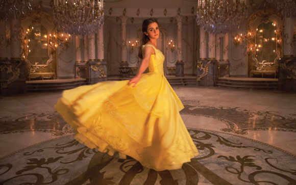 A woman in a flowing yellow ballgown twirls in a large, ornate ballroom with crystal chandeliers.