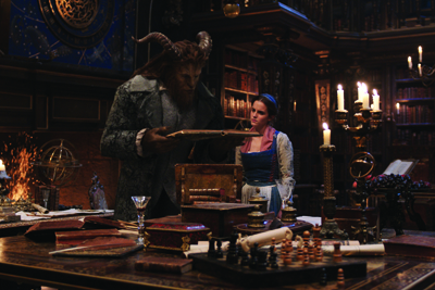 A large beast-like creature in a green coat holds a book for a woman in a blue dress in a dark library.