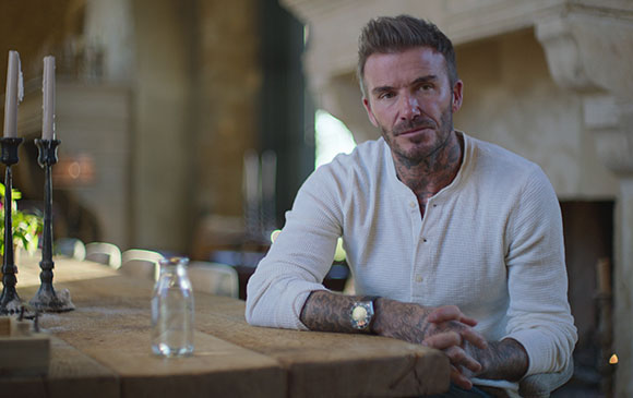 A man with tattoos and a white long-sleeve shirt sits at a rustic wooden table in a dimly lit room.