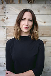 A woman with shoulder-length brown hair wearing a black turtleneck, standing before a light wood paneled wall.