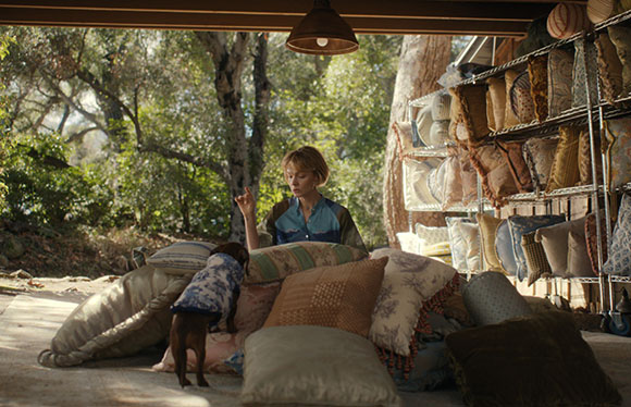 A woman sits behind a large pile of decorative pillows and cushions under a covered outdoor area.