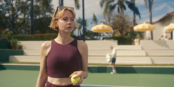 A woman in a maroon tank top holds a tennis ball on an outdoor court with palm trees and yellow umbrellas behind her.