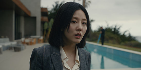 A woman with short black hair wearing a grey pinstripe blazer looks surprised near a swimming pool.