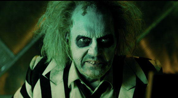 A man with pale green skin, dark sunken eyes, and messy hair wearing a black and white striped suit.