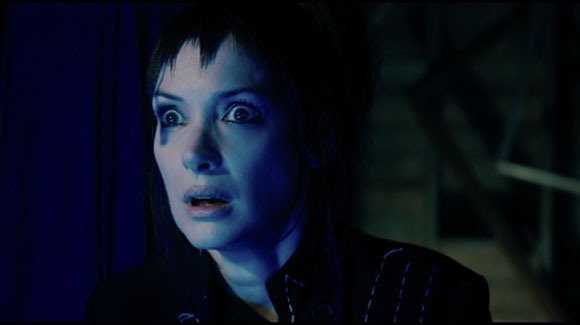 A woman with dark hair and wide eyes looks ahead with a startled expression in blue lighting.