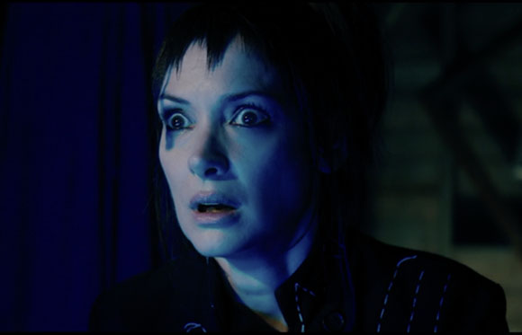 A woman with dark hair and wide eyes looks on with a shocked expression under blue light.