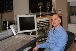 A man in a blue shirt sits at a professional audio mixing desk in a studio with awards behind him.
