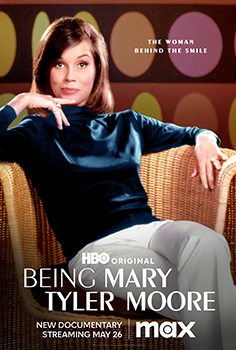 A woman in a dark turtleneck sits in a wicker chair against a background of colorful circles.
