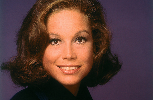 A woman with wavy brown hair and a smile looking toward the camera against a purple backdrop.