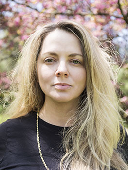 A woman with long blonde hair looks directly at the camera against a background of pink flowering trees.