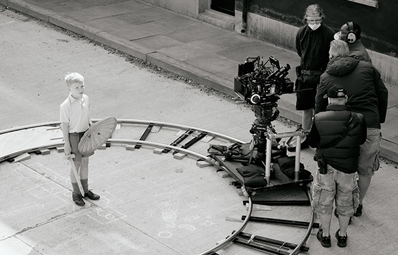 A young boy holding a prop stands inside a circular camera track being filmed by a crew on a sidewalk.