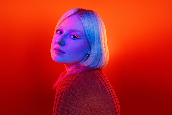 A person with blonde hair and bright orange eyeliner looking toward the camera against a red background.