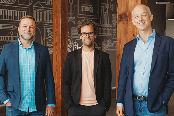 Three men in business casual attire standing in a row against a dark wall with white line art.