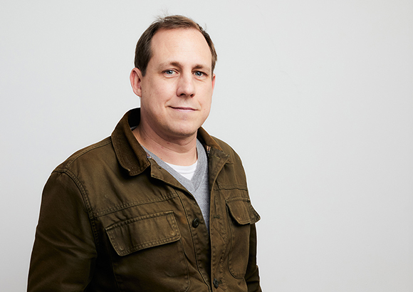 A man with short brown hair wearing a brown utility jacket over a grey shirt against a white background.