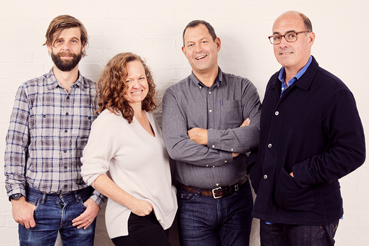 Four people, two men and two women, standing side by side against a white brick wall.