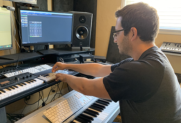 A man wearing glasses adjusts controls on a MIDI keyboard in a home studio with monitors and speakers.