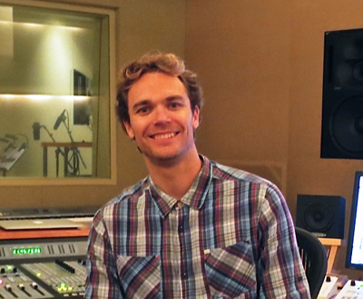 A smiling man in a plaid shirt sitting in a music recording studio.