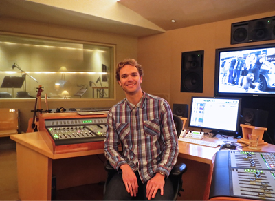 A smiling man in a plaid shirt sits at a mixing desk in a professional recording studio.