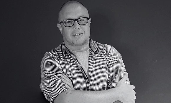 A man with glasses and a checkered shirt crosses his arms against a dark background.