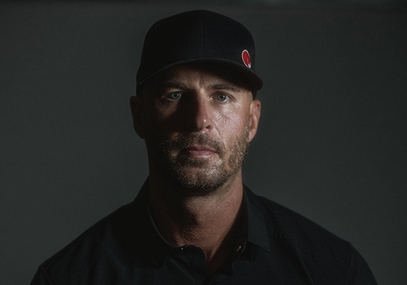 A man with a beard and a black baseball cap looks directly at the camera against a dark background.