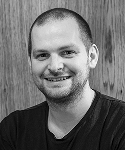 A smiling man with short hair and stubble wearing a black t-shirt against a wood-paneled background.