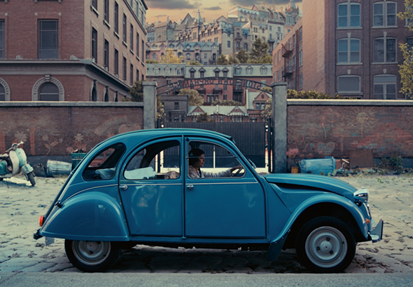 A blue vintage Citroën 2CV parked on a cobblestone street in front of a brick wall and urban buildings.