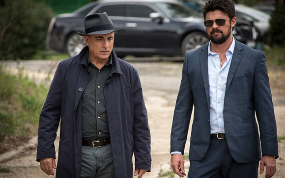 Two men in suits walk side by side on a dirt path near a parked black car.