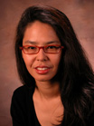 A woman with long dark hair and red-rimmed glasses smiling against a brown background.
