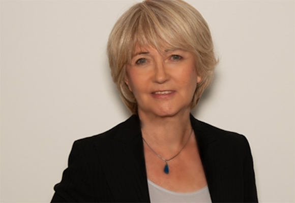 A blonde woman wearing a black blazer and a necklace with a blue pendant against a white background.