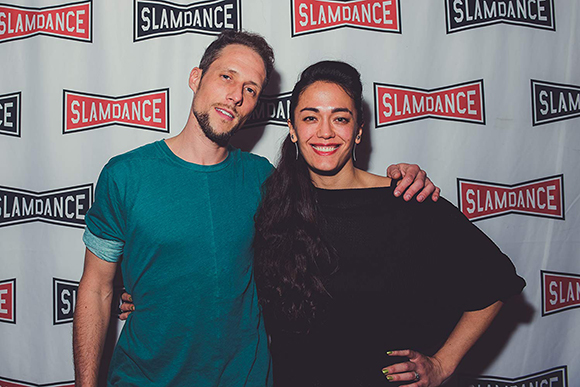 A man in a teal shirt and a woman in a black top smiling in front of a Slamdance logo backdrop.
