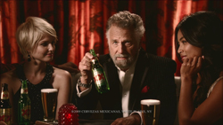 A man in a suit holds a green beer bottle while sitting between two women in a dimly lit room.