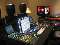 A large digital audio mixing console with multiple faders, monitors, and studio speakers in a dimly lit room.