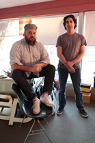 A man with a beard sits on a black chair while another man stands beside him in an office.
