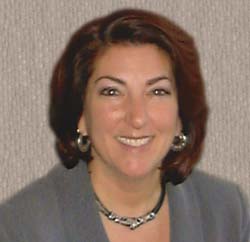 A smiling woman with shoulder-length reddish-brown hair, wearing a grey blazer and silver jewelry.