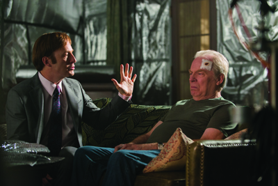 A man in a suit gestures toward a seated man with a bandage on his forehead in a dark room.