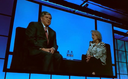 A man in a suit and a woman in a patterned blouse sit on stage against a large blue screen.