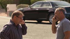 Two men stand outdoors talking on mobile phones with a dark sedan parked in the background.