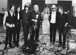 Seven musicians standing in a row in a studio setting, some holding instruments.