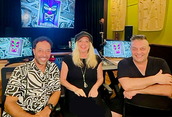 Three people sit in a music studio in front of computer monitors displaying colorful digital art.