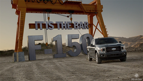 A dark Ford F-150 pickup truck parked in a dirt lot next to a large orange crane lifting massive letters.