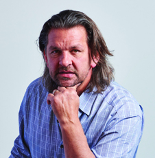 A man with shoulder-length brown hair wearing a blue plaid shirt, resting his chin on his hand.